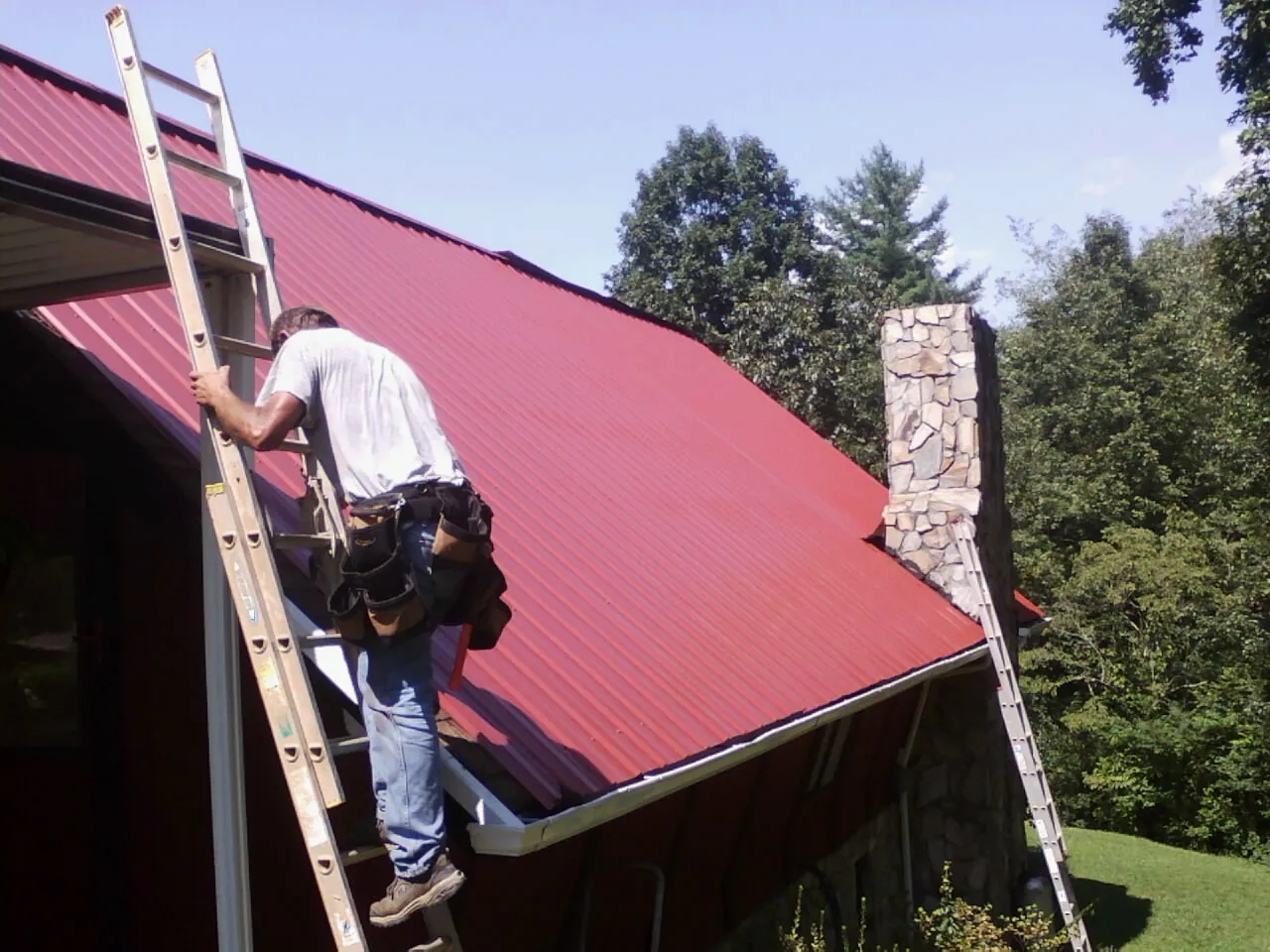 Roofer on a ladder beside a red metal roof and stone chimney — Soffit & Fascia Repair by Roofing Brothers in Trexlertown
